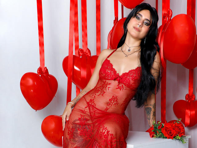 SamanthaGio in red lace lingerie, sitting with her tits visible, black hair, tattoos, red balloons, roses, and ribbon background, looking sexy, smiling, wearing a necklace.
