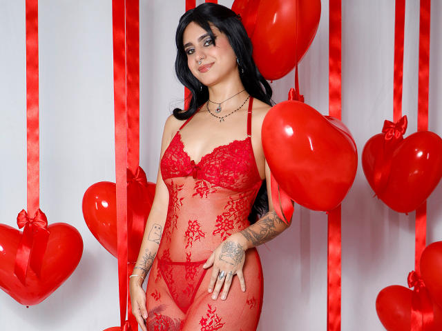 SamanthaGio with black hair, wearing red lace lingerie, tattoos on arms, holding red heart balloons, smiling, standing against white background with red ribbons. Her ass is covered.