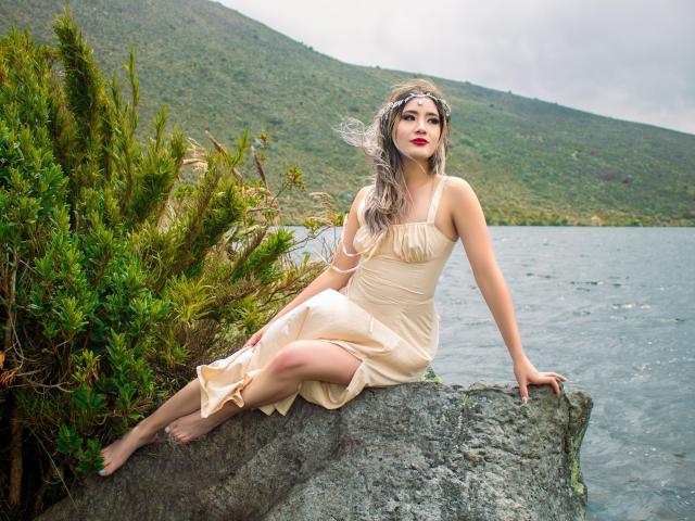 StarDustt with pale skin, red lips, and long gray hair sits on a rock, wearing a beige dress, exposing her ass, in a green, bushy, lake-side setting.