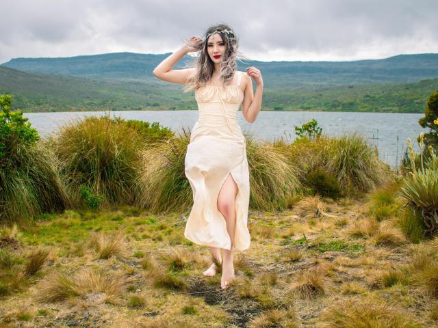 Stardustt has pale skin, long gray hair, and wears a cream dress with a thigh slit. She's barefoot, standing on grass with her hand in her hair.