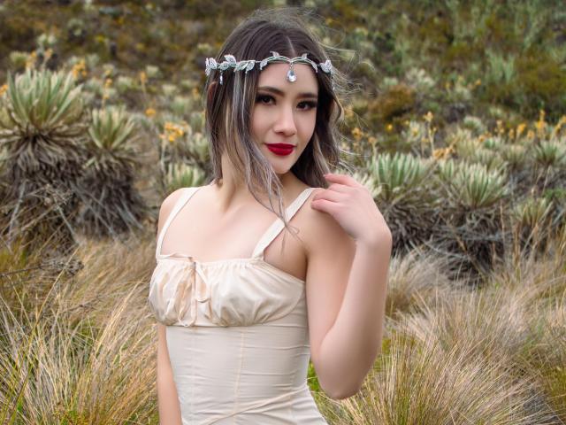 Stardustt with wavy brown hair, red lipstick, and a leaf headpiece, wearing a cream dress, touching her chin, standing in a grassy field, showing her tits.