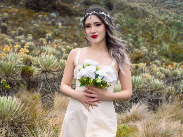 Stardustt with gray wavy hair, wearing a white dress and floral headband, holding a white rose bouquet, has red lipstick, and is standing in a grassy, rocky area.