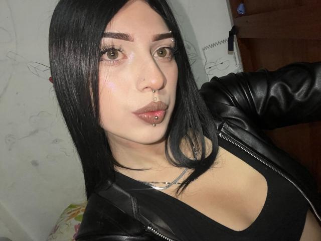 Isabella Rossi, com cabelo preto, olhos verdes, piercing no nariz e lábio, vestindo uma jaqueta de couro preta e uma blusa preta, mostrando seus pequenos seios. Ela está olhando para a câmera..