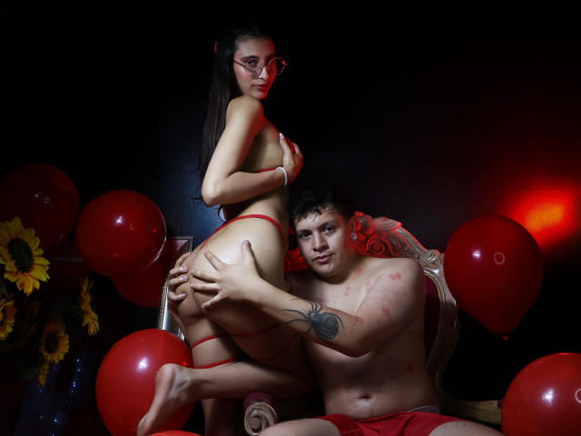 EthanNAbby with big ass, red thong, and glasses, sits on chair, while EthanNAbby with tattooed arm, red shorts, and red marks, grips her butt. Red balloons around.