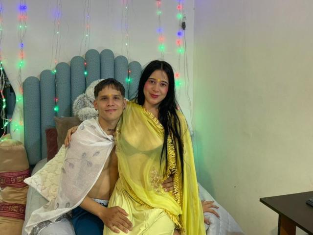 AnjaliRohan with long black hair, yellow saree, and bare shoulders, sitting on a bed next to a shirtless boy with a white cover, colorful string lights in background.