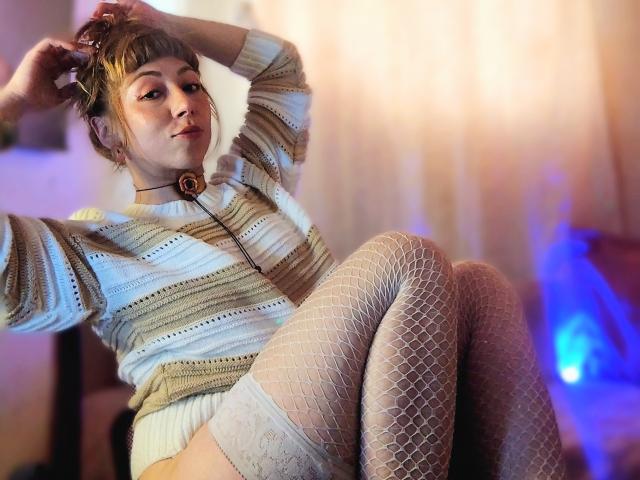 DouceKhrystine with blonde hair in a bun, wearing a striped sweater, fishnet stockings, and a choker, sitting with hands in hair, legs crossed, smiling at the camera.