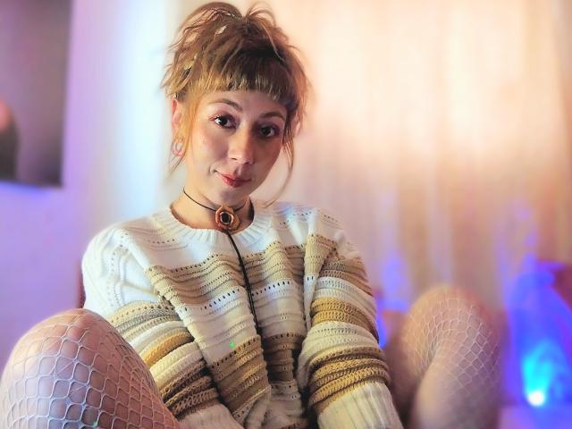 DouceKhrystine with messy brown hair, wearing a striped white knit sweater, choker with a ring, and white fishnet leggings, sitting with legs up, smiling softly.