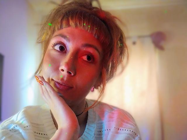 DouceKhrystine with red and green light effects on her face, pensive expression, hand on cheek, white knit top, messy blonde hair, pink earrings, soft indoor lighting, colorful highlights.
