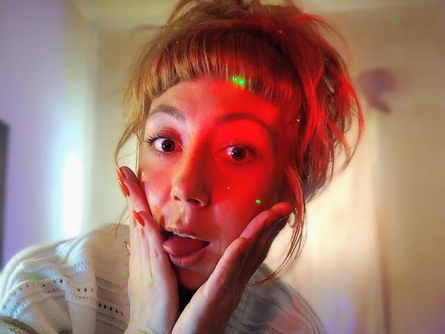 DouceKhrystine with red-lit face, hands on cheeks, tongue out, wide eyes, messy hair, and a white knit sweater. Glowing green and red light reflections on her face.