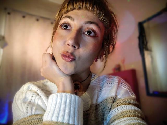 DouceKhrystine with pouty lips, big eyes, and flushed cheeks, wearing a white knitted sweater, hand on neck, brown hair in bangs, soft lighting, looking slightly up.