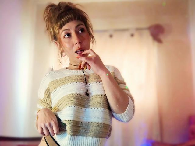 DouceKhrystine with her hand to her mouth, wearing a striped sweater, looking surprised, holding a black cord, light brown hair in a messy bun, soft light on her face.