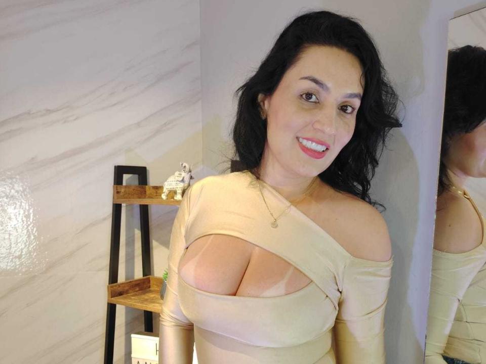 VegaRose with big tits in a beige, cut-out top, smiling at the camera, black wavy hair, pink lipstick, gold necklace, and a wooden shelf with small white figurines.