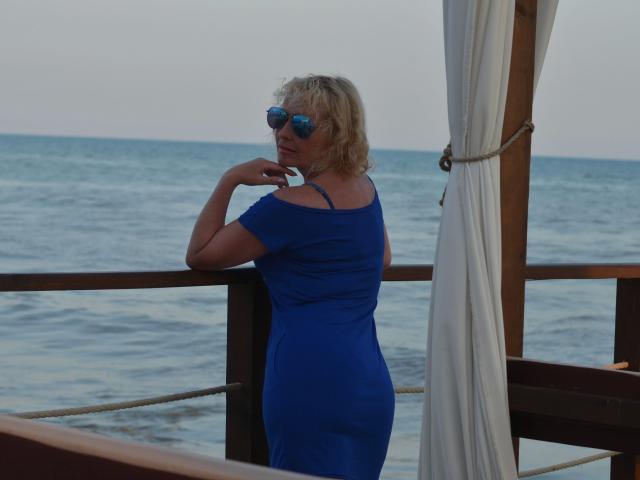 SunnySoul-hot, blonde, wearing blue dress, off-shoulder, small tits, blue sunglasses, leaning on wooden railing, white curtain, ocean view, rope, wooden structure, back to camera.
