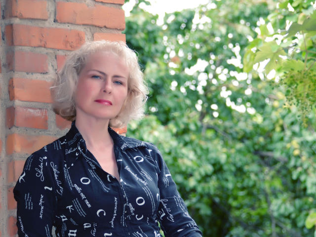 SunnySoul-hot with short blonde hair, red lipstick, and blue patterned shirt, leaning against a brick wall, green foliage in background. Her tits are small, she's attractive.