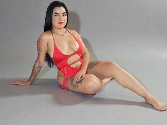 HannaStarSx with long black hair, red cut-out one-piece, tattoos on arms and thigh, sitting on gray floor, legs spread, red lips, looking at camera, red nails.