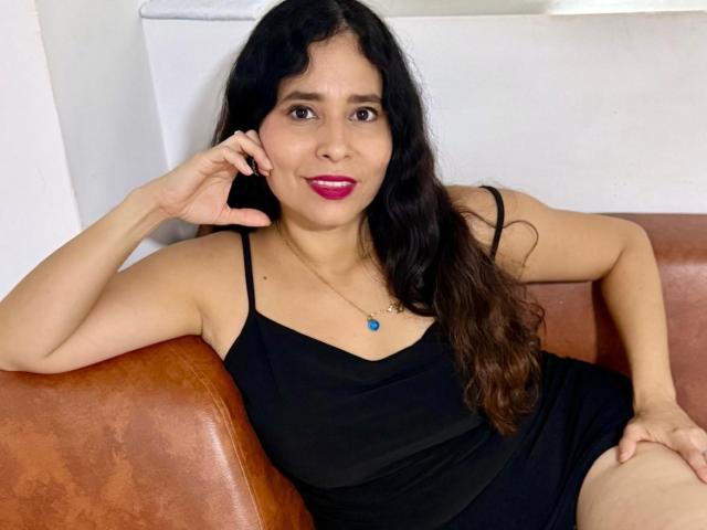 MiaEverhart with black wavy hair, pink lipstick, black dress, blue necklace, sitting on brown leather chair, right arm resting on armrest, smiling, right hand near ear.