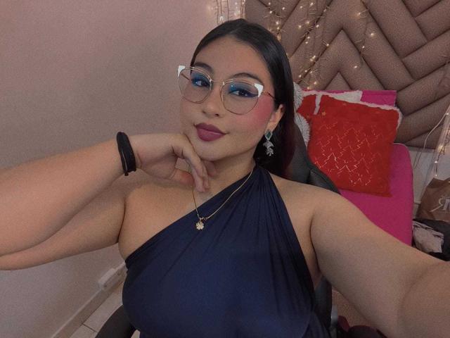 HelueJamil with glasses, dark hair, navy halter dress, pink lipstick, necklace, black bracelet, resting chin on hand, smiling, red pillow in background.