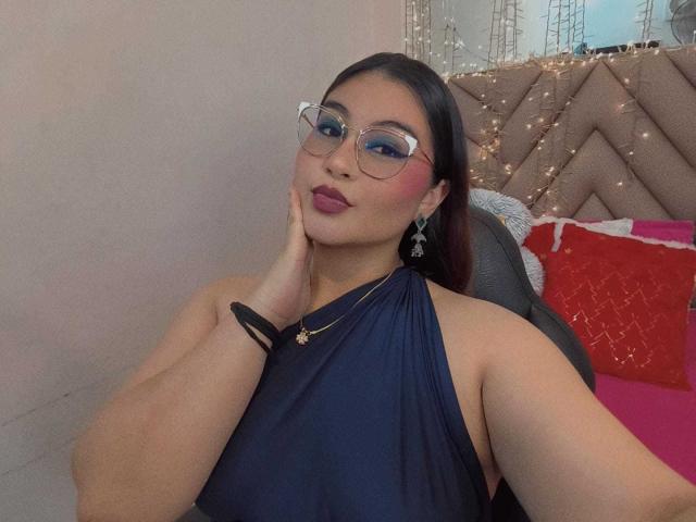 HelueJamil with dark hair, glasses, blue dress, hand on cheek, pink lipstick, necklace, and earrings, sitting in chair, looking at camera, slight smile. Boobs visible under dress.