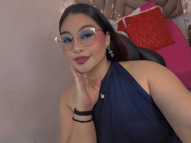 HelueJamil with glasses, blue eye makeup, dark lipstick, navy halter top, hand on cheek, black bracelet, earrings, and a flower pin, sitting in a chair.