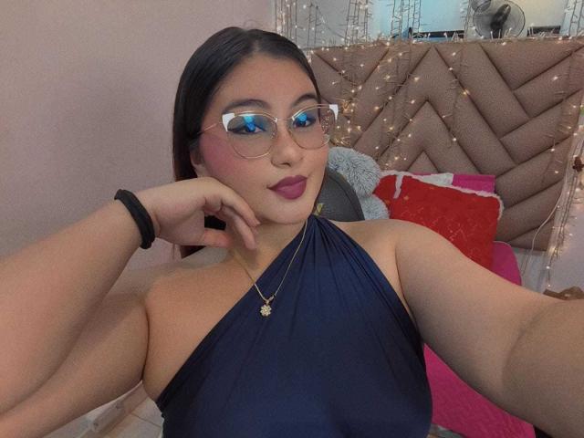 HelueJamil with blue eyes, glasses, dark lipstick, navy halter top, necklace, black wristband, resting chin on hand, smiling, medium tits, dark hair, pink wall, bed with pillows.