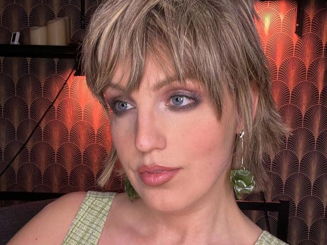 CharleneAngel-hot has short blonde hair, blue eyes, and is wearing green earrings with leaf patterns. She's got pink lipstick and a slight smile, looking directly at the camera.