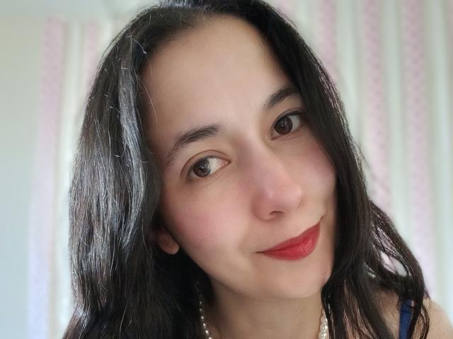 AnneStone's face with dark, wavy hair, brown eyes, light skin, and red lipstick. She's smiling slightly, wearing a pearl necklace, and has small, subtle earrings.