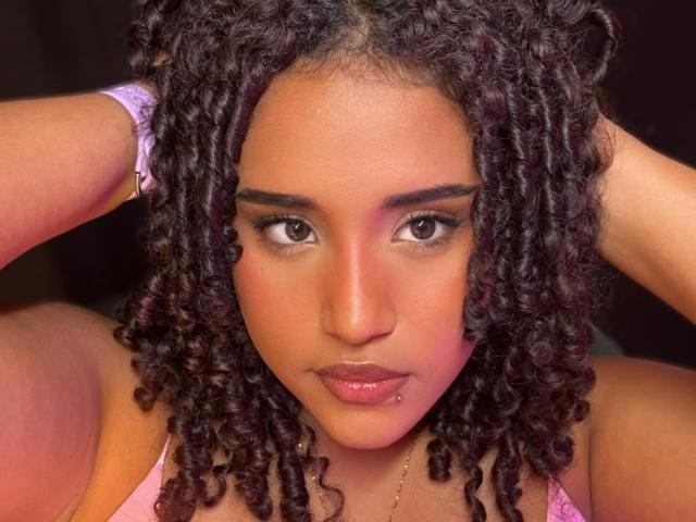 MelodyDubois has curly brown hair, tan skin, and a lip piercing. She's looking at the camera with hands in her hair, wearing a pink top.