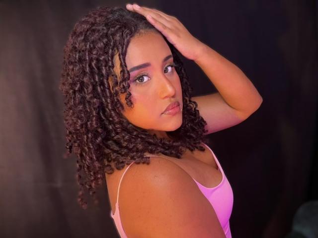 MelodyDubois with curly brown hair, wearing a pink tank top, has her hand on her head, looking over her shoulder, showing her medium-sized tits and a serious expression.