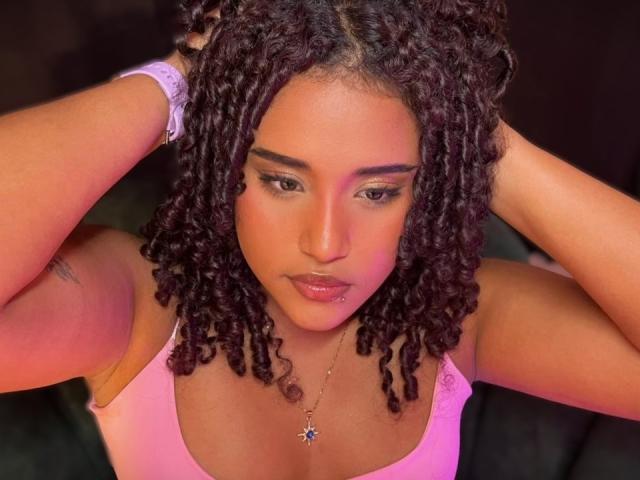 MelodyDubois with curly dark brown hair, pink tank top, hands in hair, looking down, glossy pink lips, necklace with star pendant, purple wristband, warm orange lighting.