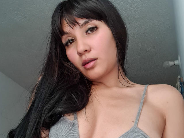 NaylaGrey with long black hair, bangs, and gray spaghetti top, showing small tits, pink lips, and a nose ring, looking at the camera with a slight smile.