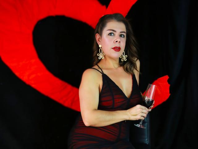 EmiliaMilf with red lipstick, black sheer dress, and gold earrings, holding a glass of red wine, standing against a black background with a large red heart.