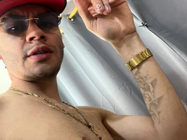 NastyX69 with a red cap, sunglasses, and gold watch, shirtless, showing a tattoo on his arm, holding his fingers up. NastyX69's medium skin and slight stubble are visible.