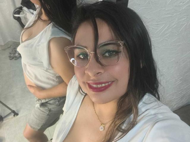 CamiilaBabe with glasses, red lipstick, and white shirt, smiling at camera, showing tits in mirror selfie, standing in front of another CamiilaBabe with white tank top and jeans.