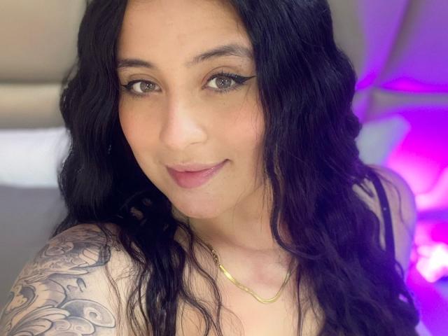 AnnyBentleii has long black wavy hair, brown eyes, and a subtle smile. She's wearing a gold necklace, showing a tattoo on her left shoulder. Boobs are partially visible.