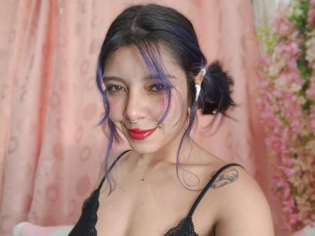 DarckVelvet with purple hair, red lipstick, black lace bra, nose ring, smiling, tattoo on right shoulder, pink curtain, and pink flowers in the background. Boobs visible.