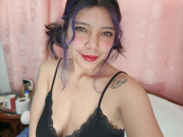 DarckVelvet has purple-dyed hair, a nose ring, and red lipstick, wearing a black lace bra, showing her tits, with tattoos on her right shoulder and left arm.