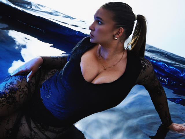 MissKuromi with large tits, wearing a black lace dress, leaning back in blue water, dark hair in a ponytail, gold earrings, and a necklace, looking to the side.