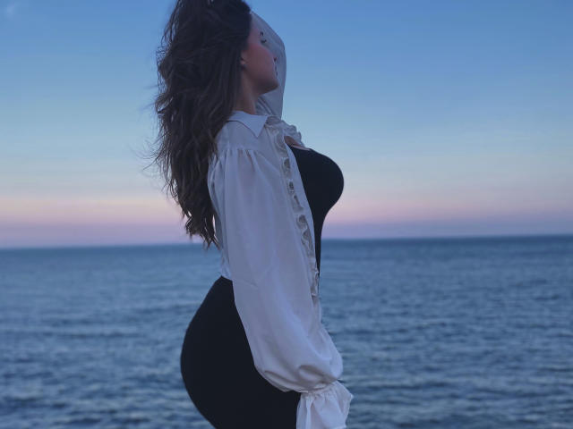 MissKuromi with curly brown hair, facing side, wearing a white shirt over black dress, highlighting her big ass and small tits, standing by a calm blue ocean at dusk.