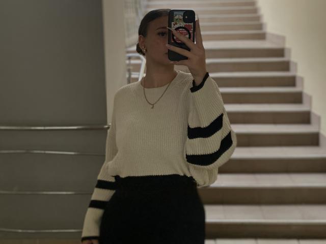 MissKuromi with dark hair in a bun, wearing a white knit sweater with black stripes and black pants, taking a selfie with a phone, showing a small gold necklace.