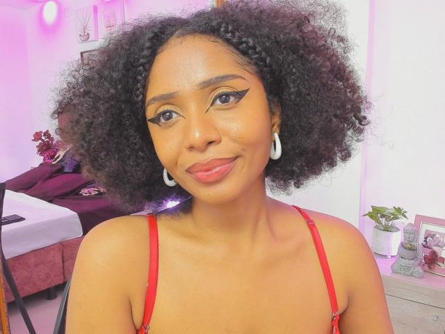 MeganWlliams with curly black hair, wearing red lingerie, has medium brown skin, white hoop earrings, and red lipstick. She's smiling with winged eyeliner and pinkish lighting.
