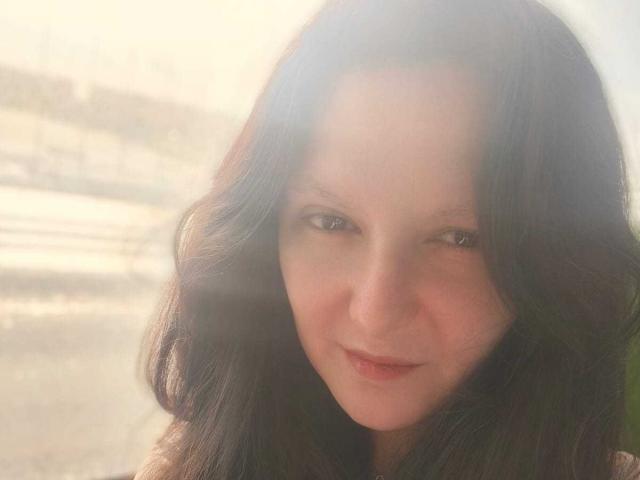 AmethystCharm has wavy dark brown hair, light skin, and a subtle smile with her eyes slightly squinted. She's looking directly at the camera with a soft, gentle expression.