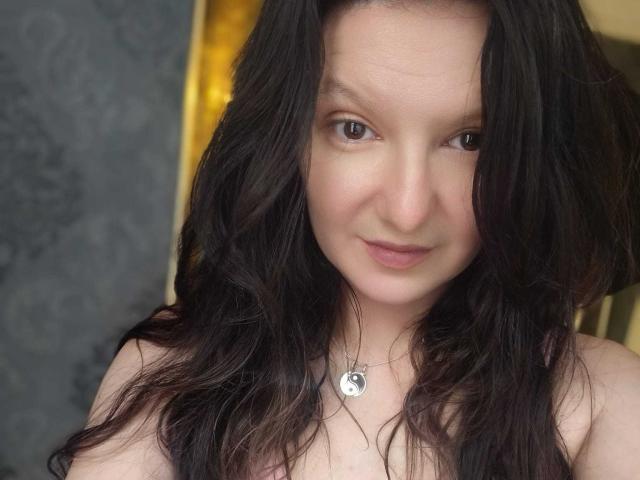 AmethystCharm has wavy dark brown hair, light skin, and is wearing a necklace with a silver circular pendant. She's smiling slightly with brown eyes and no visible clothing.