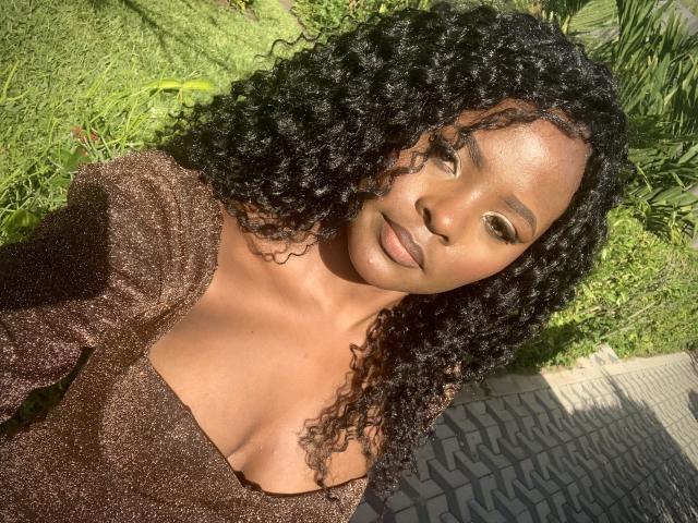 Close-up photo of SexyBadriya. She has dark, curly hair and smooth, medium-brown skin. She's wearing a sparkly, brown, low-cut top that reveals her cleavage. Her eyes are slightly upcast, and she's looking at the camera with a relaxed expression. The sunlight casts a natural glow on her face. The background includes green grass and a patterned, gray brick wall. Her makeup is subtle, with defined eyebrows and a natural lip color. The image has a warm, natural light.