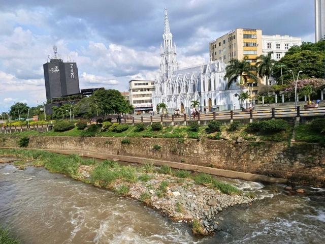 White gothic-style church with a tall spire, situated near a muddy river with grassy banks, surrounded by buildings, palm trees, and a road with a wooden fence.