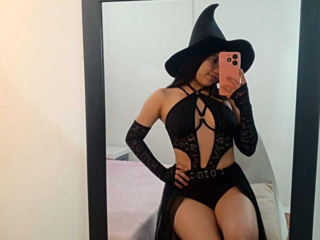 ChannelStark in a black witch outfit with a hat, lace gloves, and a revealing halter top, showing her tits and ass, taking a mirror selfie with a pink phone.