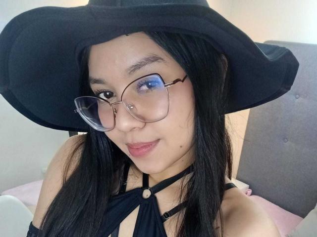 ChannelStark with black hat, glasses, and blue eye makeup, showing off her tits in a black strappy top. She's got long black hair and a cute nose piercing.