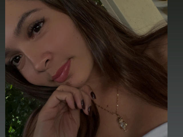Close-up of Gabrielafoxxy with long brown hair, light brown skin, and subtle makeup. She has full pink lips and dark, slightly arched eyebrows. Her right hand with black-painted nails rests on her cheek. She wears a gold necklace with a small pendant. The image is slightly shadowed, highlighting her facial features. Gabrielafoxxy's expression is calm and slightly playful. The background is blurred, focusing on her face.