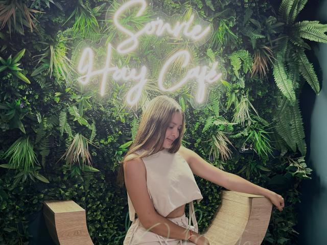 Gabrielafoxxy with long blonde hair, small tits, wearing a white sleeveless crop top and white skirt, sitting on a wooden chair, neon sign above, green plant wall behind.