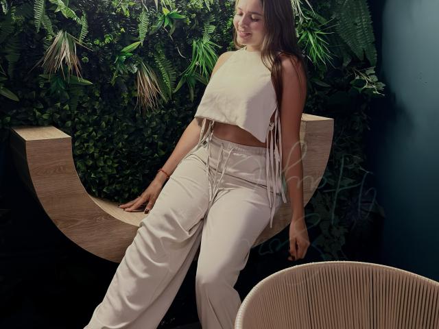 Gabrielafoxxy sits on a wooden, curved bench. She's wearing a white, sleeveless crop top with fringes and high-waisted, loose-fitting white pants. Her long brown hair is straight and she has a slight smile. The background is dense green foliage. She's positioned slightly to the right with her left hand on the bench and right hand resting on her lap. A wicker chair is in the bottom right corner. The lighting is soft, creating subtle shadows.