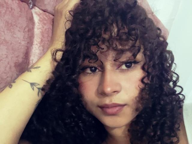 Sofia, com cabelos pretos encaracolados, olhos castanhos e batom rosa, segurando a cabeça com uma tatuagem no braço. Seu rosto tem um leve semblante de descontentamento, olhando para o lado..
